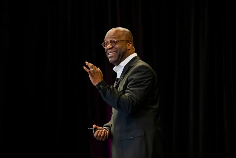 A speaker gesturing with a smile while addressing an audience. He is wearing a black suit and glasses, standing in front of a dark backdrop that transitions to shades of purple.