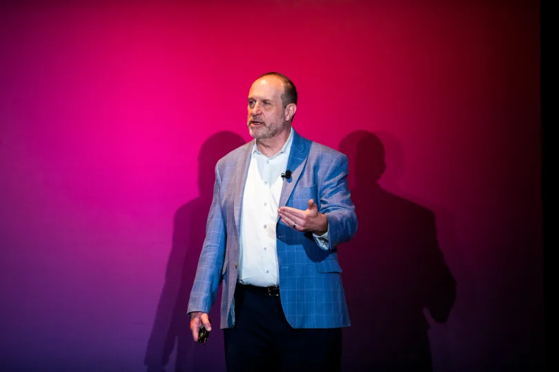 A speaker stands on stage in front of a vibrant pink backdrop, gesturing with one hand while holding a microphone in the other. The individual appears engaged and animated as they address an audience. The scene captures the energy of a live presentation, highlighting the speaker's interaction with the audience.