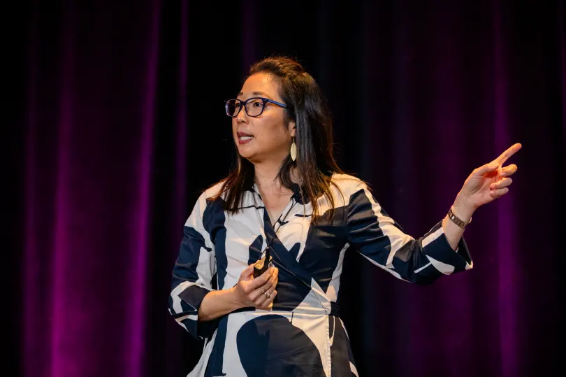 A woman with glasses is speaking passionately on stage, gesturing with her right hand to emphasize her points. She is wearing a black and white patterned dress. The background features deep purple curtains, creating a vibrant stage setting.