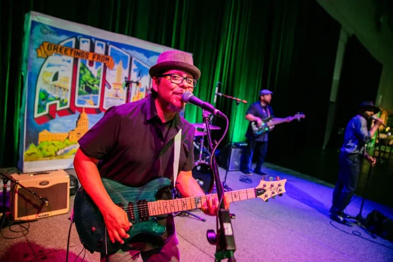 A musician playing a blue electric guitar and singing into a microphone, performing on stage. Behind him is a backdrop featuring a colorful mural that reads 'Greetings from Austin.' Two other band members can be seen in the background, one playing the bass guitar and another preparing to sing. The stage is illuminated with vibrant lighting, creating a lively atmosphere.