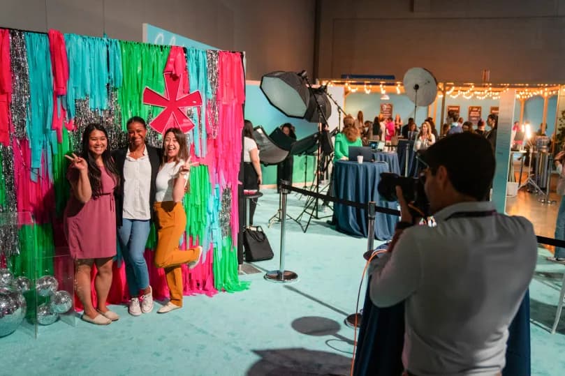 Three women are posing for a photo in front of a vibrant photo backdrop made of colorful streamers in shades of pink, green, and silver. One woman is making a peace sign while the others smile. In the background, a camera is raised by a photographer capturing the moment, and there are additional event attendees mingling and tables set up with various items.