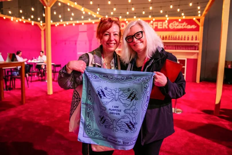 Two women smile while holding a blue bandana that reads 'I AM PURPOSE DRIVEN.' They stand in a venue decorated with string lights and a pink backdrop. In the background, there are tables and chairs, indicating a social or event setting.