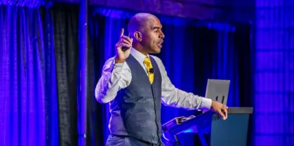 A speaker passionately presenting on stage with one hand raised, wearing a suit and tie. The backdrop features blue lighting and curtains, creating an engaging atmosphere. A laptop is positioned on a podium beside the speaker.