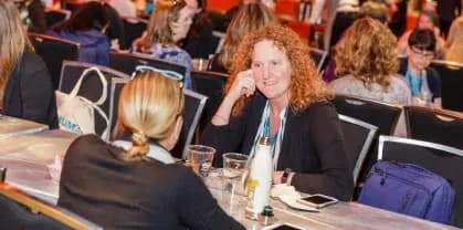 A woman with curly red hair is smiling while engaged in a conversation with another person at a conference. They are seated at a table, and the atmosphere is lively with other attendees visible in the background, some with bags and drinks on their tables. The setting suggests a network or discussion environment typical of conference events.