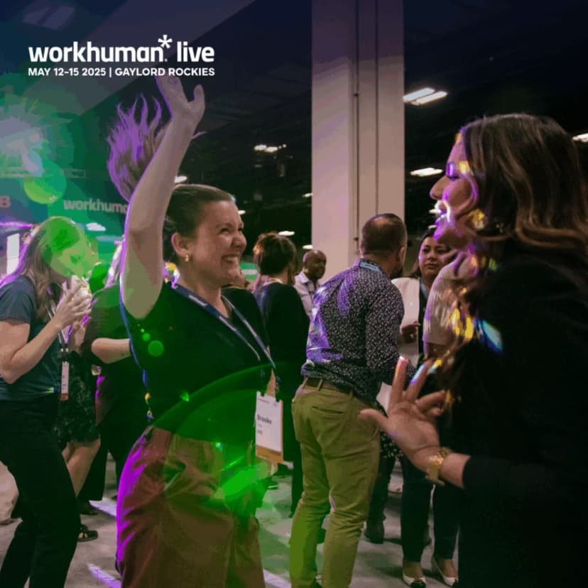 A lively scene from the Workhuman Live conference, featuring attendees enjoying the event. One woman, smiling and animated, is dancing with her arm raised, while another woman engages with her. Various attendees are seen in the background, contributing to a vibrant atmosphere.