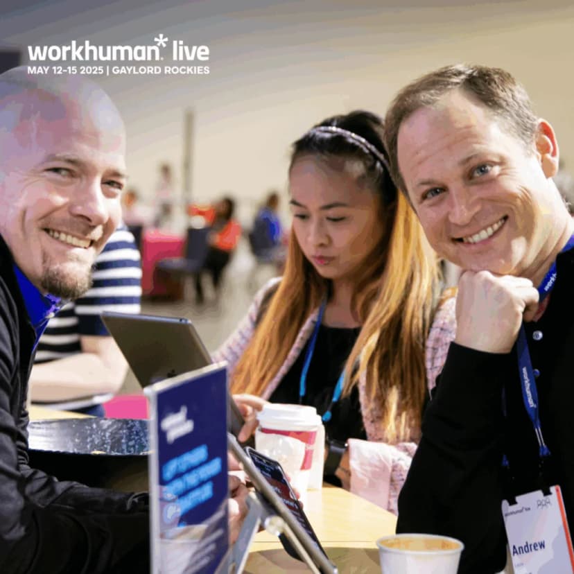 A man and a woman are seated at a table with electronic devices, appearing to engage in discussion at the Workhuman Live event. The man on the left has a shaved head and is smiling, while the woman next to him is focused on her tablet, displaying long hair and wearing a black top. In the foreground, a coffee cup is visible, and a name tag featuring 'Andrew' is placed on the table.