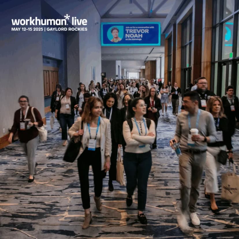 A crowded hallway at the Workhuman Live conference shows a diverse group of attendees walking together. The atmosphere is vibrant and engaged, with many individuals wearing name badges and carrying bags. A large screen in the background features an advertisement for a conversation with Trevor Noah, scheduled for May 12-15, 2025, at the Gaylord Rockies venue. The setting is modern, with colorful carpet design and bright overhead lighting, reflecting a dynamic conference environment.