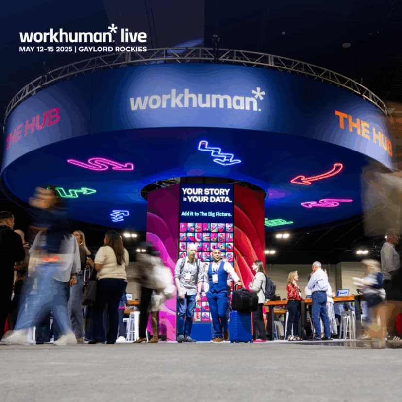 A vibrant display at the Workhuman Live conference featuring the 'The Hub' exhibit. The booth showcases text reading 'YOUR STORY IS YOUR DATA. Add it to The Big Picture,' surrounded by attendees engaging with one another, some carrying luggage. Neon arrows and designs decorate the ceiling, adding to the energetic atmosphere of the setting.