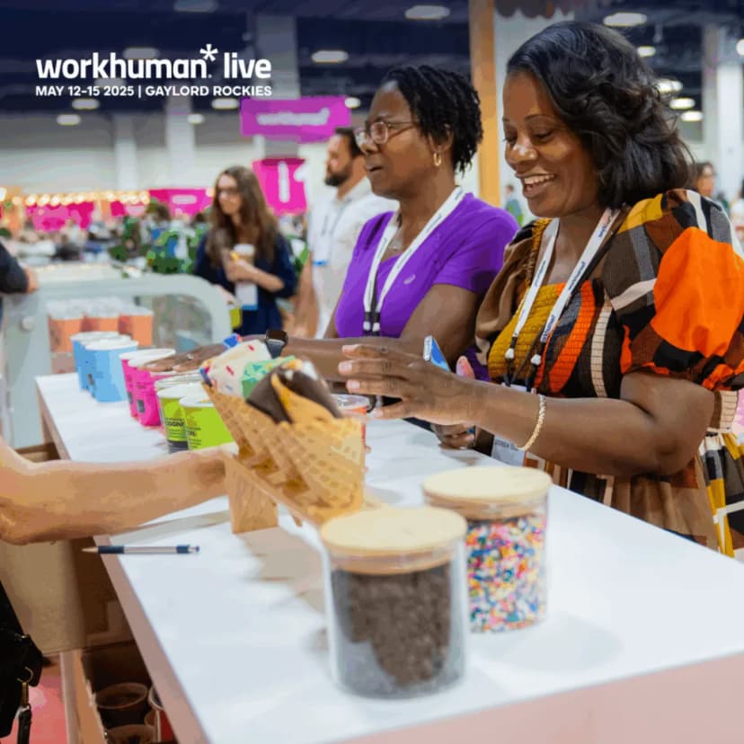 Two women are engaged in a cheerful conversation while selecting ice cream from a display at the Workhuman Live event. The booth features colorful containers and toppings, and both women are smiling as they look at the offerings.