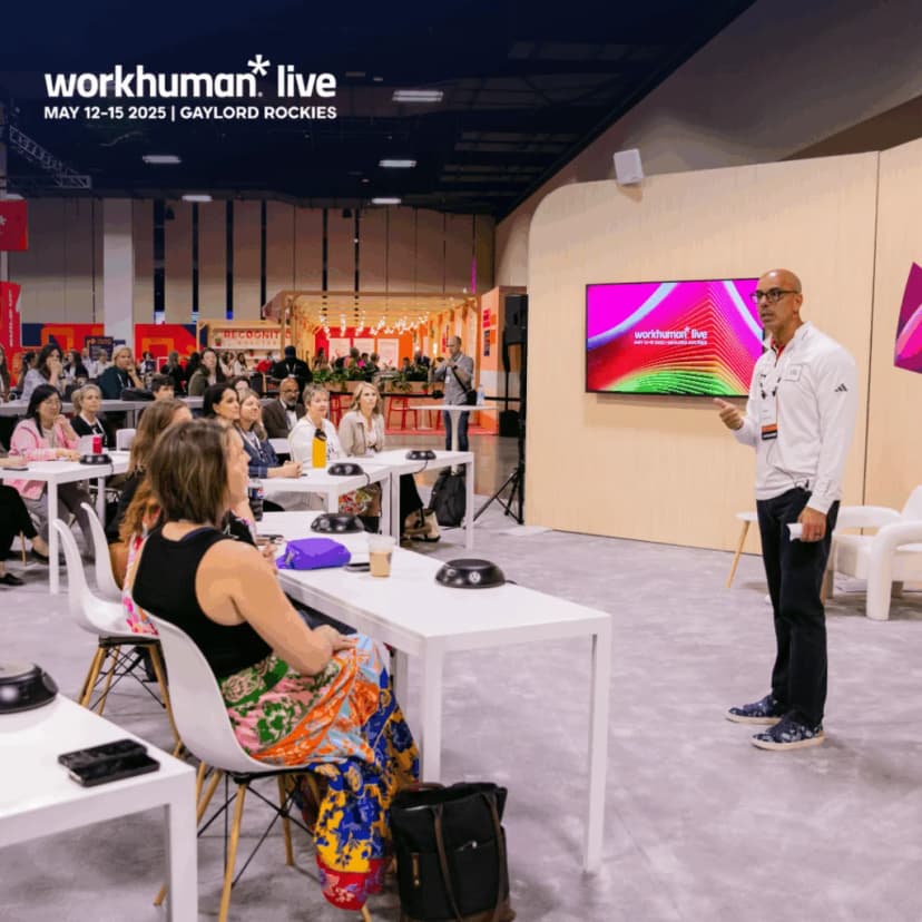 A speaker presenting at the Workhuman Live event, engaging with an audience seated at tables. The setting includes a large screen displaying the Workhuman Live logo and colorful graphics. Attendees, mostly women, are focused on the presentation, with a few taking notes or drinks in hand. The ambiance is professional and collaborative, set in a large conference space.