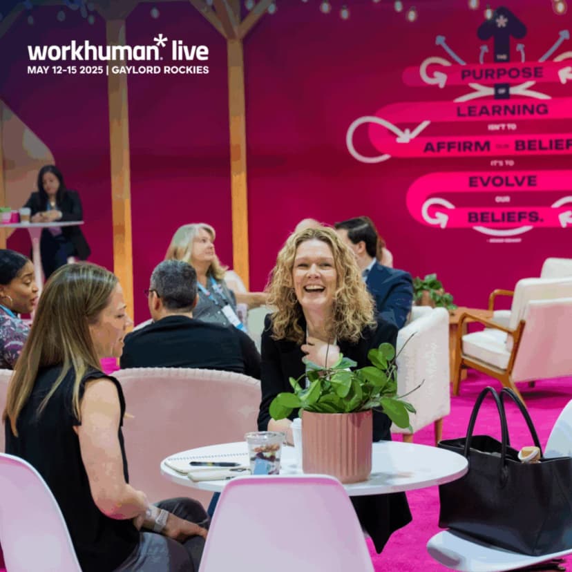 A lively scene from the Workhuman Live conference, featuring a group of attendees engaging in conversation at a round table. One woman with curly hair is laughing while seated next to another woman, with a potted plant on their table. In the background, other conference participants are visible, with a bright pink backdrop displaying the text: 'Purpose isn't to affirm beliefs, it's to evolve our beliefs.'