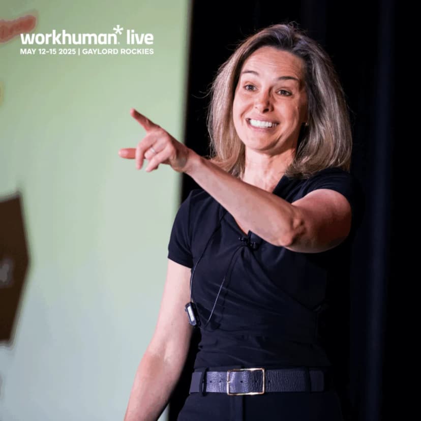 A speaker on stage enthusiastically gestures while addressing an audience at the Workhuman Live conference. The speaker has shoulder-length brown hair and is wearing a black top. The overall atmosphere is engaging and dynamic, emphasizing an interactive presentation.