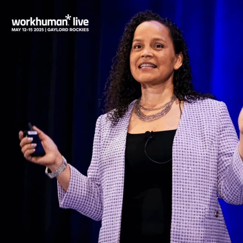 A speaker gesturing with both hands while addressing an audience, wearing a black top and a patterned light gray blazer.