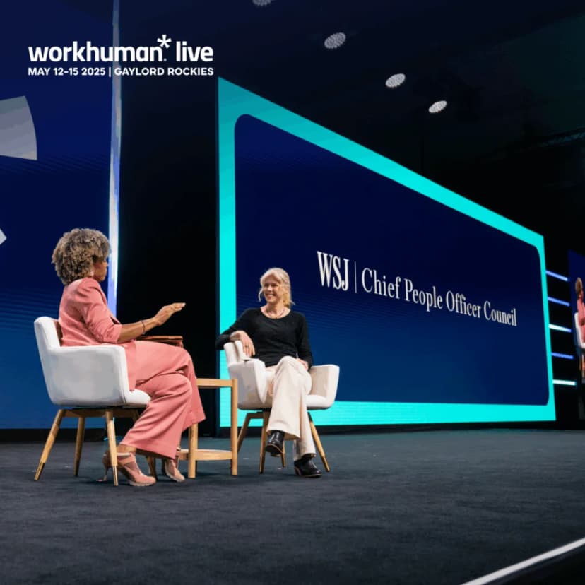 A live panel discussion at the Workhuman Live event features two women seated in chairs. One woman, dressed in pink, gestures while speaking, and the other, in a black top, listens attentively. A large screen behind them displays the text 'WSJ Chief People Officer Council,' indicating the topic of discussion. The event is set against a professional conference backdrop, with branding for Workhuman Live visible at the top.