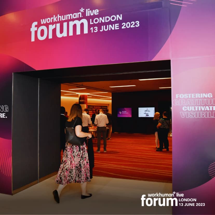 A woman in a patterned dress walks through an entrance to the Workhuman Live Forum in London, dated June 13, 2023. The entrance is adorned with vibrant signage that reads 'workhuman live forum' along with the date and phrases like 'FOSTERING GRATITUDE' and 'CULTIVATING VISIBILITY.' Inside, attendees are gathered, engaging with the event activities.