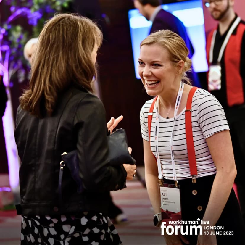 A smiling woman wearing a striped shirt and red suspenders engages in conversation with another woman in a black jacket at the Workhuman Live forum in London. The event details, including date, are displayed at the bottom of the image. Background figures add to the lively atmosphere of the conference.