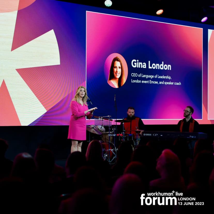 A woman in a pink dress stands on stage speaking into a microphone during the Workhuman Live event in London. Behind her is a large screen displaying her name, 'Gina London,' along with her title as the CEO of Language of Leadership and details about her role as an event emcee and speaker coach. In the foreground, two musicians play instruments, adding to the dynamic atmosphere of the event, which is attended by an audience visible in the lower part of the image.