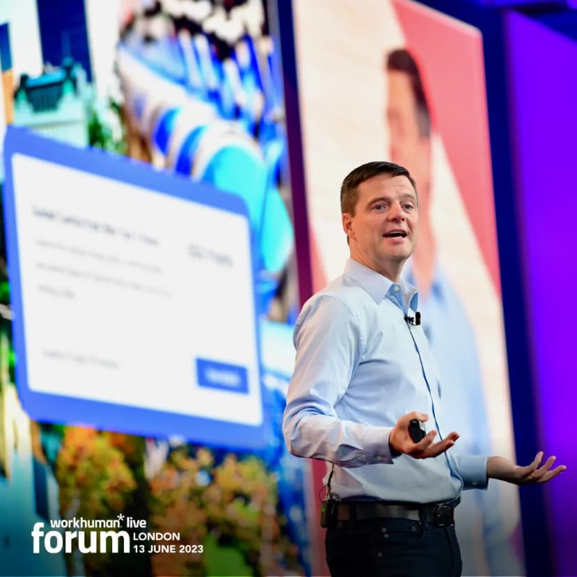 A speaker gesturing with both hands during a presentation at the Workhuman Live Forum in London on June 13, 2023. The background features colorful visuals, including a large screen displaying a digital interface.