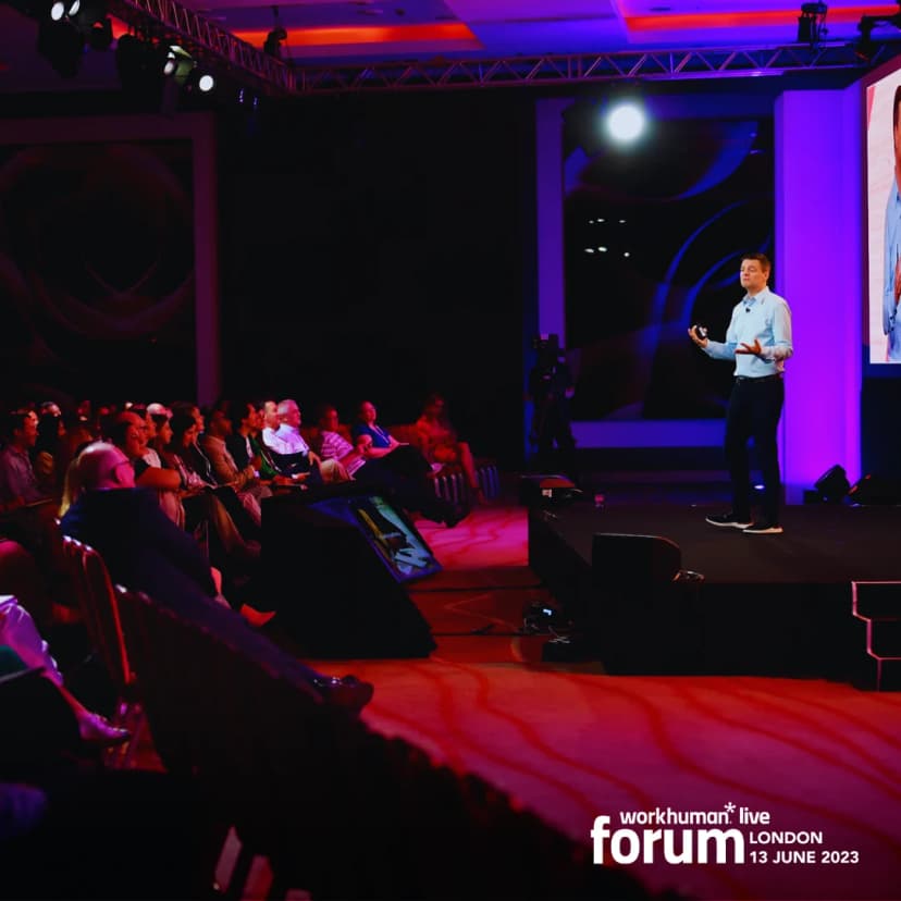 A speaker stands on stage at the Workhuman Live forum in London, engaging an audience seated in front of him. The background features a large screen displaying visuals related to the event. The scene is lit with colorful lights, creating an energetic atmosphere.