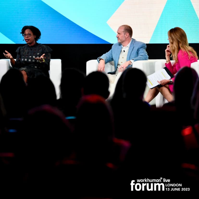 A diverse panel of speakers is engaging the audience at the Workhuman Live Forum in London on June 13, 2023. One speaker, a woman wearing glasses and a black outfit, is animatedly discussing a point, while a man in a light blue blazer listens attentively beside her. A woman in a pink outfit is taking notes and appears to be thoughtfully considering the discussion. The audience is partially visible in the foreground.