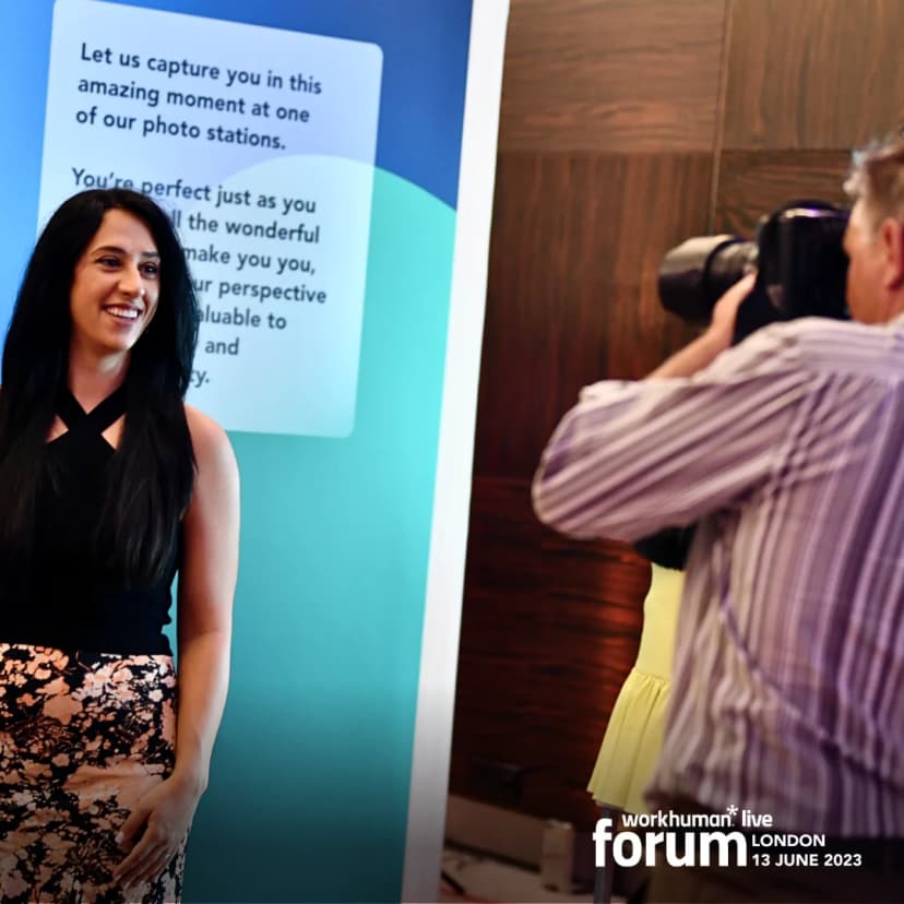 A woman smiles while posing for a photo at a Workhuman event, standing in front of a backdrop that includes a sign with text encouraging self-appreciation and capturing memorable moments. A photographer is taking her picture, and the setting appears vibrant and celebratory, with additional event elements in the background.