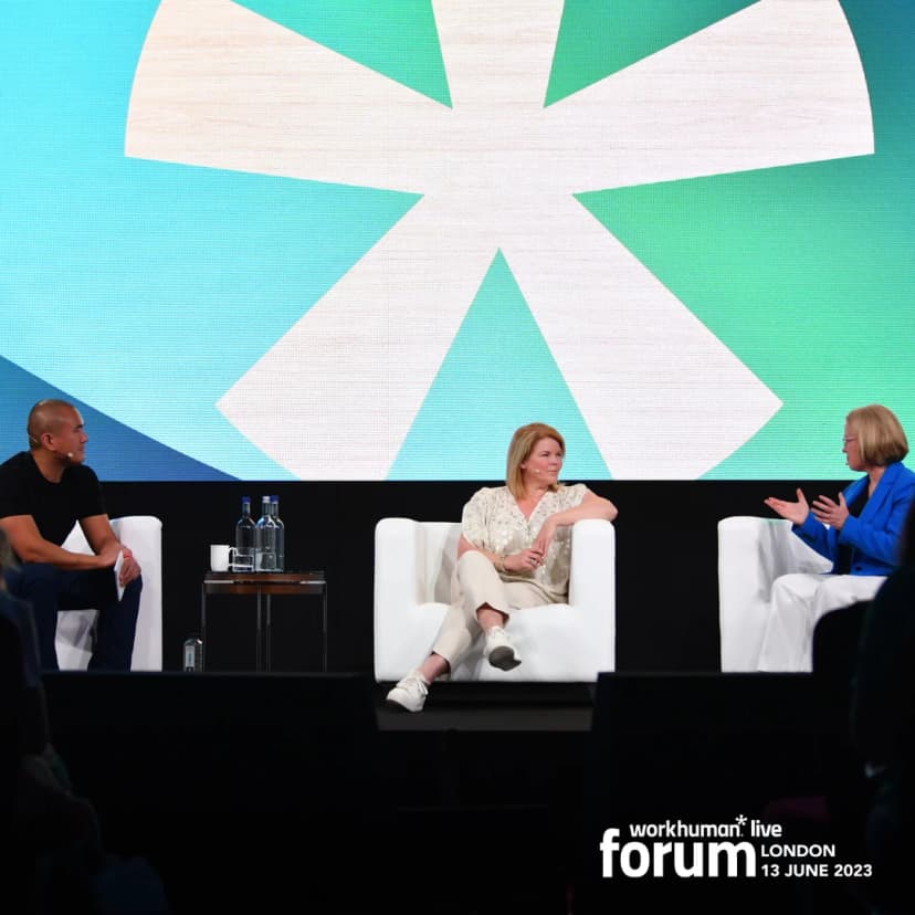 A panel discussion at the Workhuman Live forum in London on June 13, 2023, features three speakers seated on white armchairs against a colorful backdrop with a prominent Workhuman logo. One speaker is a man in a black shirt and blue jeans, while the other two speakers are women, one wearing a patterned blouse and the other in a blue jacket. A water bottle and glasses are placed on a small table next to them, creating an engaging atmosphere for the audience.