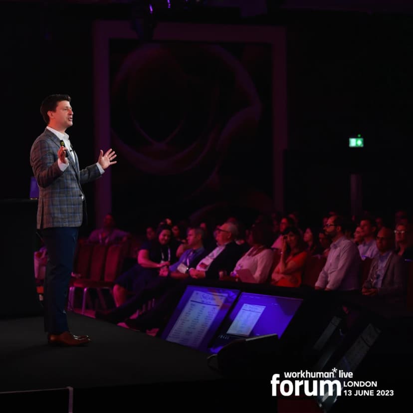 A speaker stands on stage at the Workhuman Live forum in London, addressing an audience seated in the background. The presenter gestures with open arms, wearing a plaid jacket. The backdrop features a large image of a rose, while two screens display data and content relevant to the event.