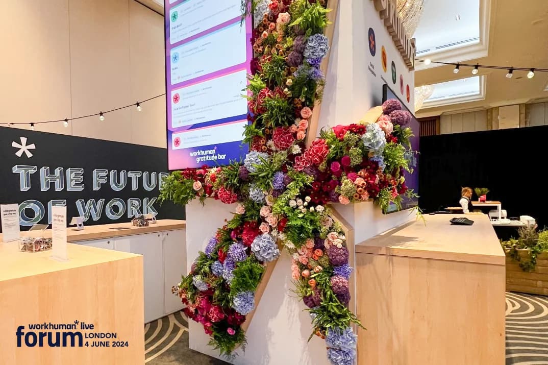 A floral arrangement in vibrant colors forming an oversized 'X' shape, displayed at the Workhuman Gratitude Bar. Behind it, a digital screen features several recognition messages. To the side, a sign reads 'The Future of Work.' The setting includes wooden surfaces and the decor is complemented by ambient lighting.