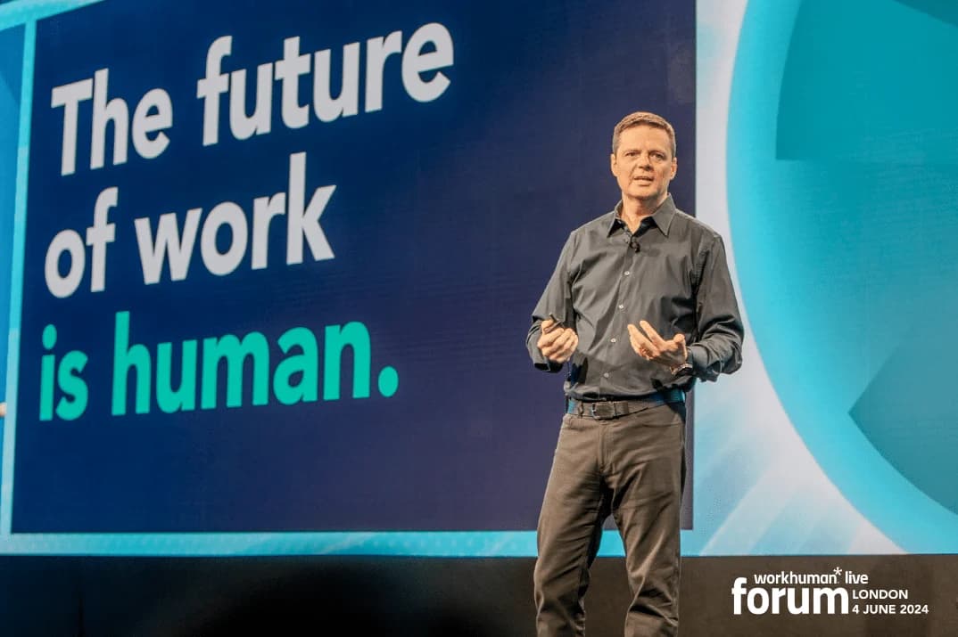A speaker standing on stage at the Workhuman Live Forum in London, presenting to an audience. The background features the text 'The future of work is human,' prominently displayed on a large screen. The speaker appears to be engaged with the audience while gesturing with their hands.
