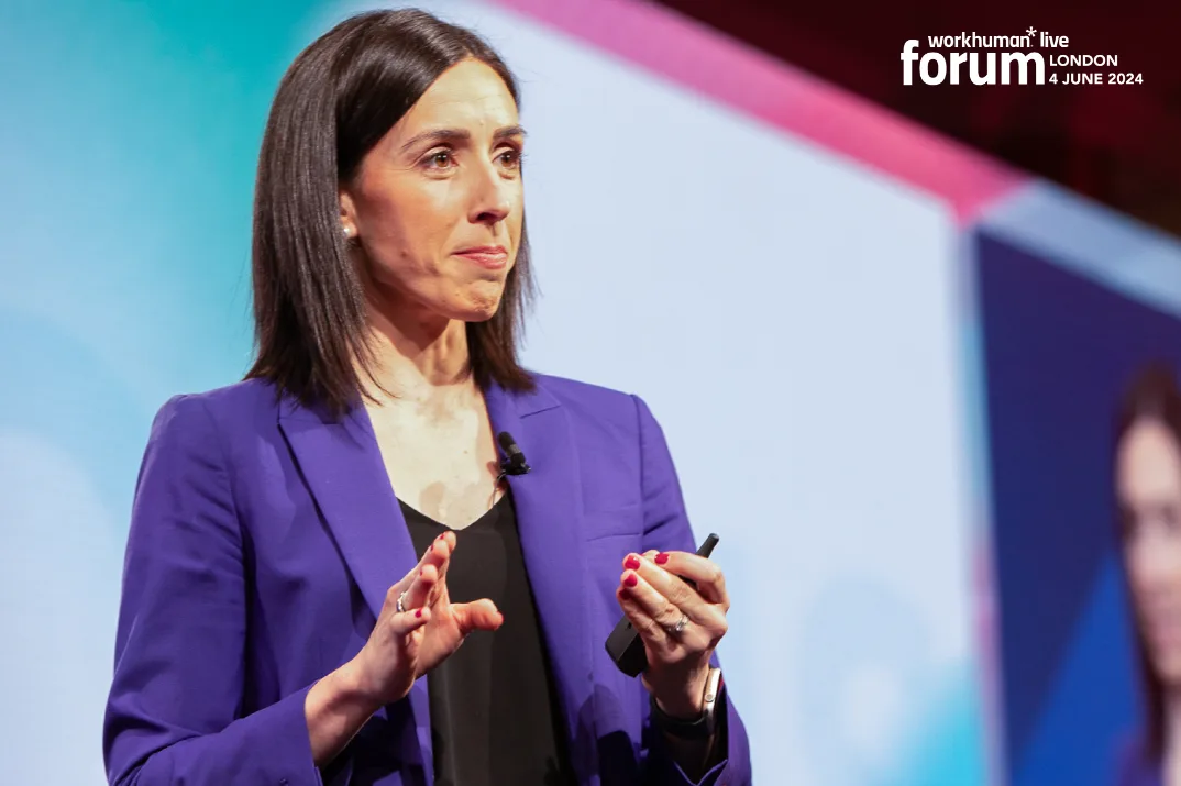 A woman in a blue blazer is speaking at the Workhuman Live forum event in London on June 4, 2024. She appears engaged and is using hand gestures while holding a presentation remote. A colorful background is visible, adding to the conference atmosphere, and the event branding can be seen in the top right corner of the image.