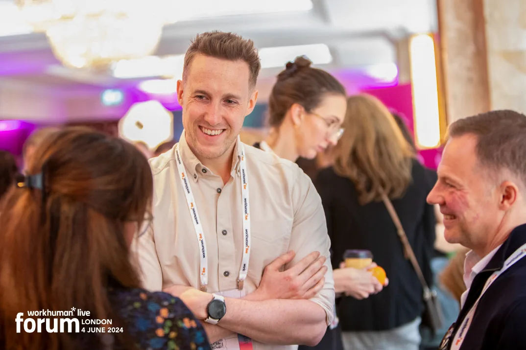 A group of attendees engaging in conversation at the Workhuman Live conference in London. One man is smiling and interacting with a woman, while others are visible in the background. The setting has bright, colorful lighting and a professional atmosphere.