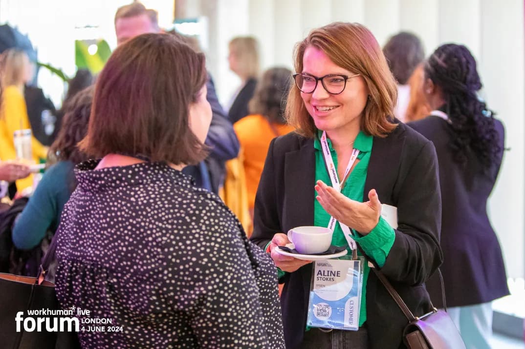A smiling woman with brown hair and glasses is engaged in conversation with another woman while holding a small cup. She is wearing a black blazer over a green top and has a name badge that reads 'Aline Stokes'. In the background, other attendees are visible, creating a vibrant conference atmosphere.