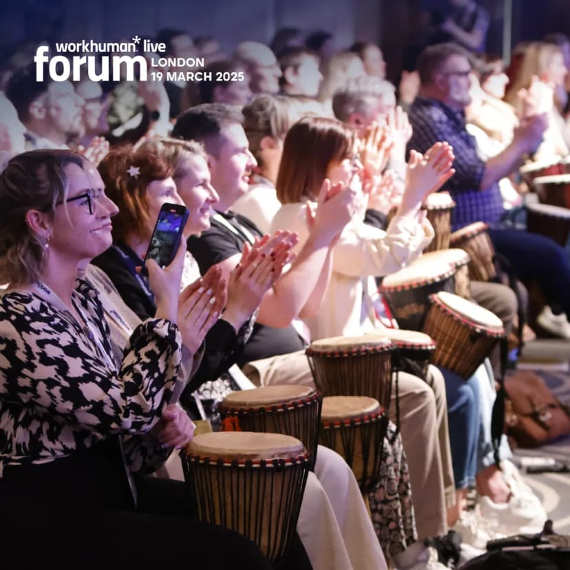 A lively audience at the Workhuman Live Forum in London, featuring attendees clapping enthusiastically. In the foreground, a woman with glasses smiles while holding a phone. The audience, wearing a variety of outfits, is seated with hand drums in front of them, showing a vibrant and engaged atmosphere.