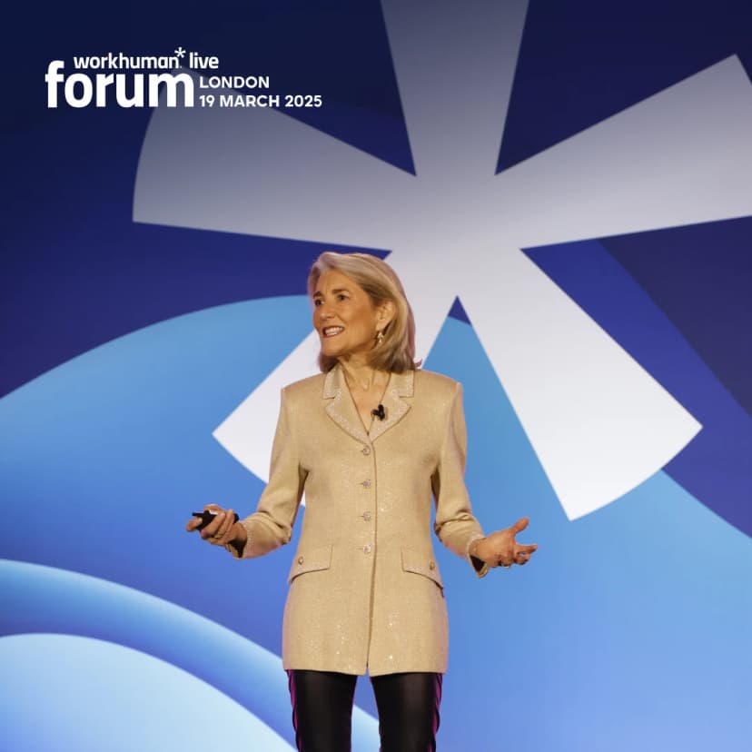 A speaker wearing a sparkling beige blazer stands on stage, gesturing with a microphone in hand. Behind her is a blue background with a large white star-like shape. She appears engaged and confident while addressing the audience.