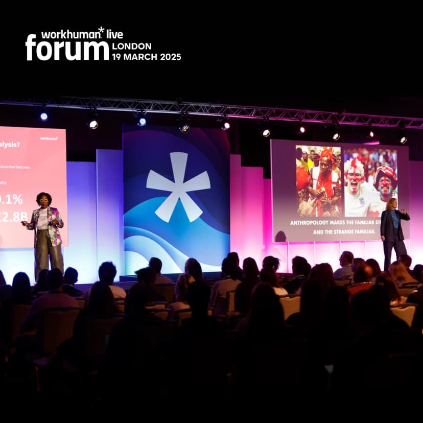A speaker presenting at the Workhuman Live forum in London on March 19, 2025, stands on stage wearing a colorful outfit while another speaker gestures with their hand to emphasize a point. The backdrop features a large logo and vibrant decorations, with a slide displaying the concept 'Anthropology makes the familiar strange and the strange familiar.' An audience can be seen seated in front, engaged in the presentation.