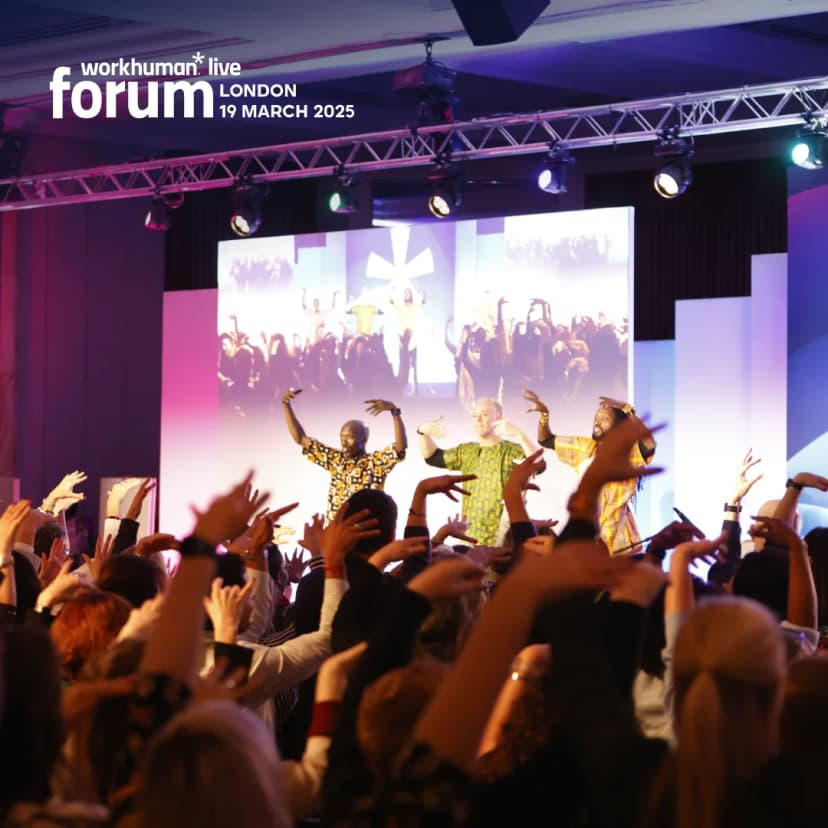 A lively scene from the Workhuman Live Forum in London on March 19, 2025, shows two performers engaging with an enthusiastic audience, who are raising their hands. The stage features a vibrant backdrop and large screens displaying energetic visuals, creating an atmosphere of participation and excitement.