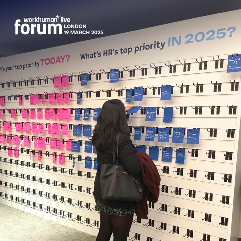 A participant at the Workhuman Live forum in London on March 19, 2025, is seen interacting with a display board filled with colorful sticky notes. The wall showcases questions about HR priorities, with large texts asking, 'What's your top priority TODAY? and 'What's HR's top priority IN 2025?' The board features pink and blue notes that communicate various topics, including 'Inclusion' and 'Equity,' while the individual wearing a dark blazer is reaching for a blue note.