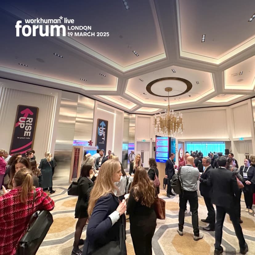 A bustling gathering at the Workhuman Live Forum in London, featuring a diverse group of attendees engaged in conversation. The venue showcases elegant decor and displays, with prominent signs reading 'RISE UP' and 'LIFT' against the backdrop of the event date, March 19, 2025. The atmosphere is dynamic, suggesting a professional networking event.