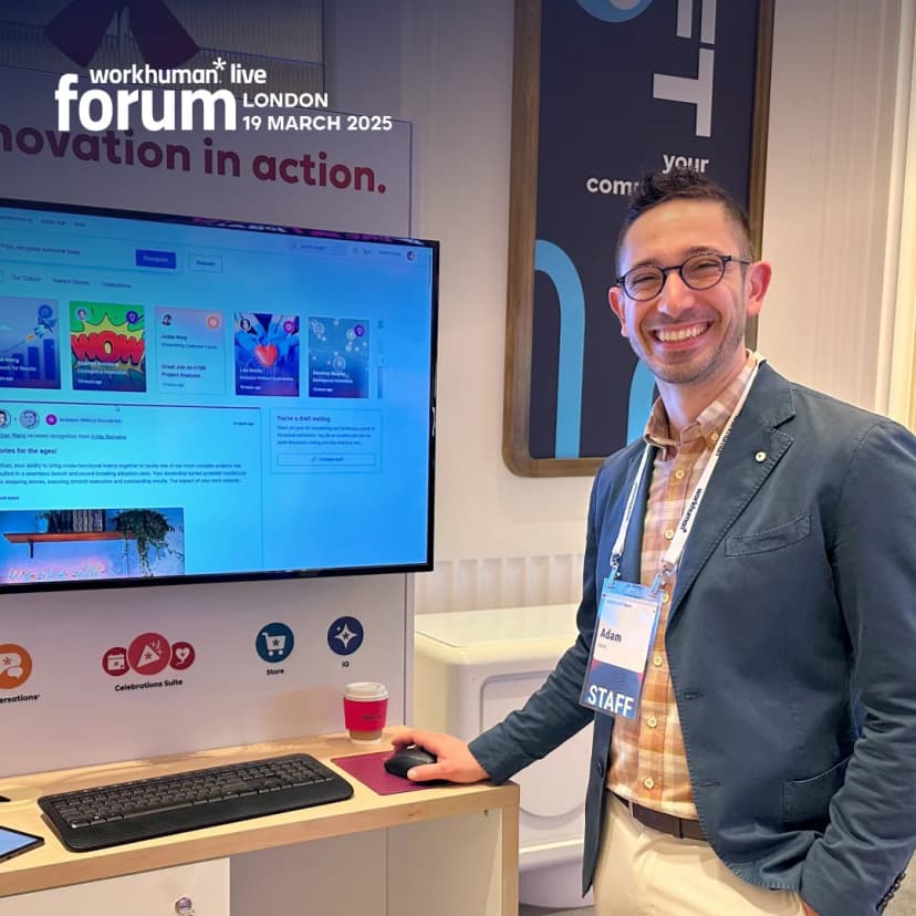A smiling man named Adam stands in front of a computer monitor displaying various colorful recognition messages and resources related to work culture, at the Workhuman Live Forum in London. The text on the screen mentions concepts like 'innovation in action.' He is wearing a blazer over a plaid shirt, and a badge labeled 'STAFF.' The setting appears to be an exhibition space designed for engaging conversations and recognition highlights.