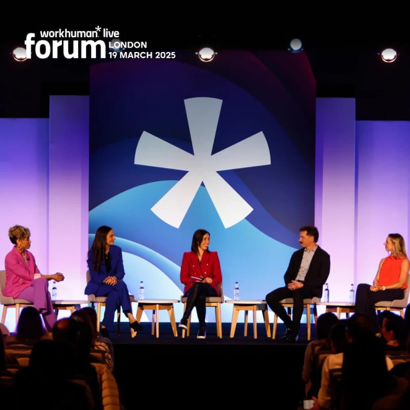 A panel of five speakers, including two women dressed in bright outfits and a man in a dark suit, is seated on stage at a Workhuman event. The audience is partially visible in front of the stage.