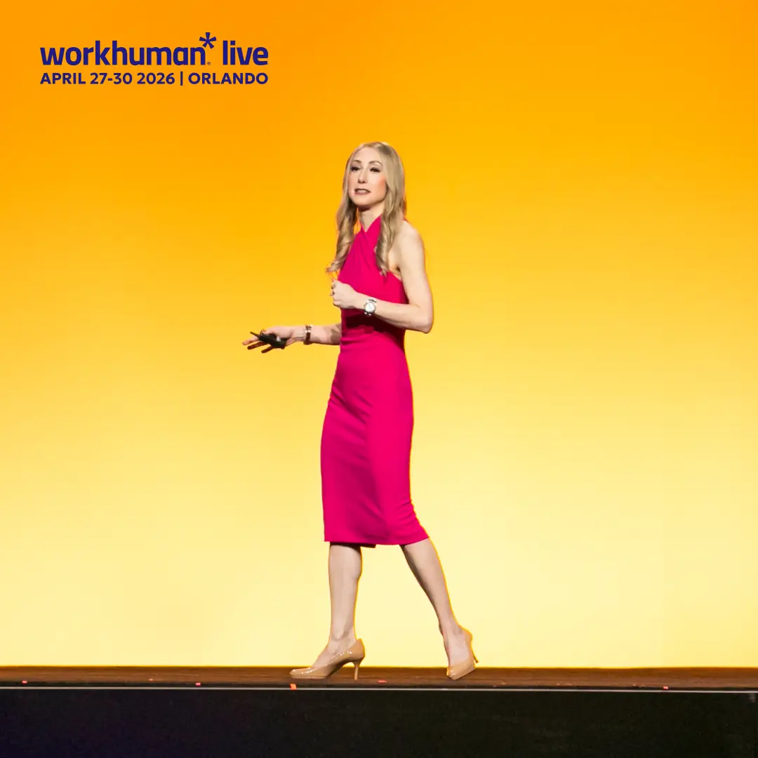 A woman in a pink dress is speaking on stage with a bright orange backdrop at the Workhuman Live event. The text at the top of the image reads "workhuman live APRIL 27-30 2026 | ORLANDO." Her hand gestures suggest she is engaged in delivering a presentation.