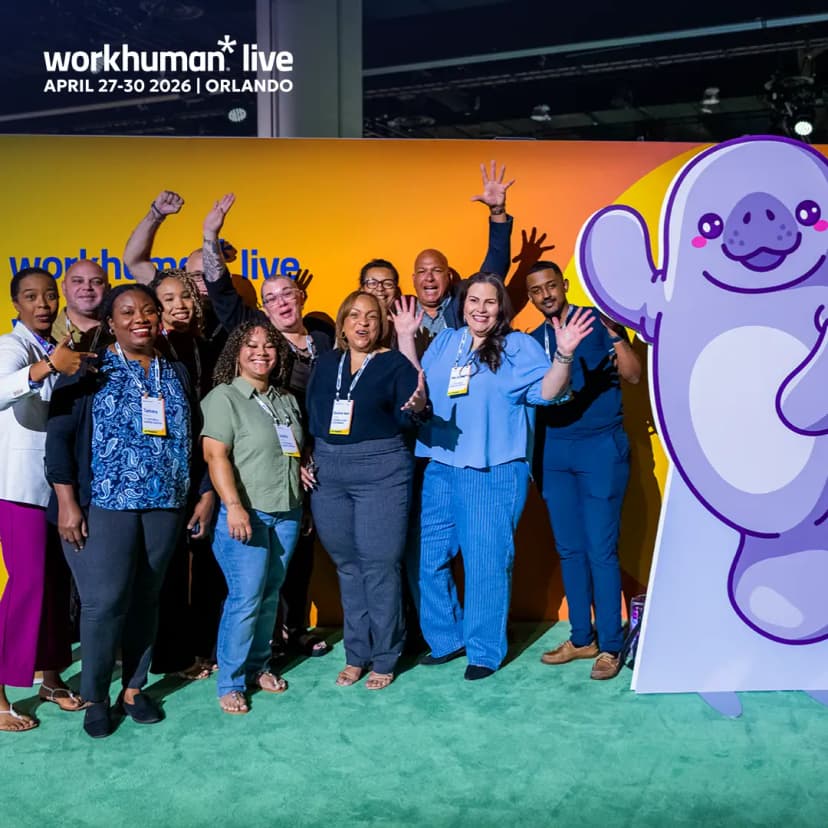 A group of diverse individuals is gathered for a photo in front of a colorful backdrop that reads "workhuman live" along with the dates "APRIL 27-30 2026" and "ORLANDO." They are smiling and waving, displaying a sense of excitement and camaraderie. To the right of the group, there is a playful illustration of a cartoon character resembling a friendly animal. The setting appears vibrant and welcoming, indicative of a conference or event atmosphere.