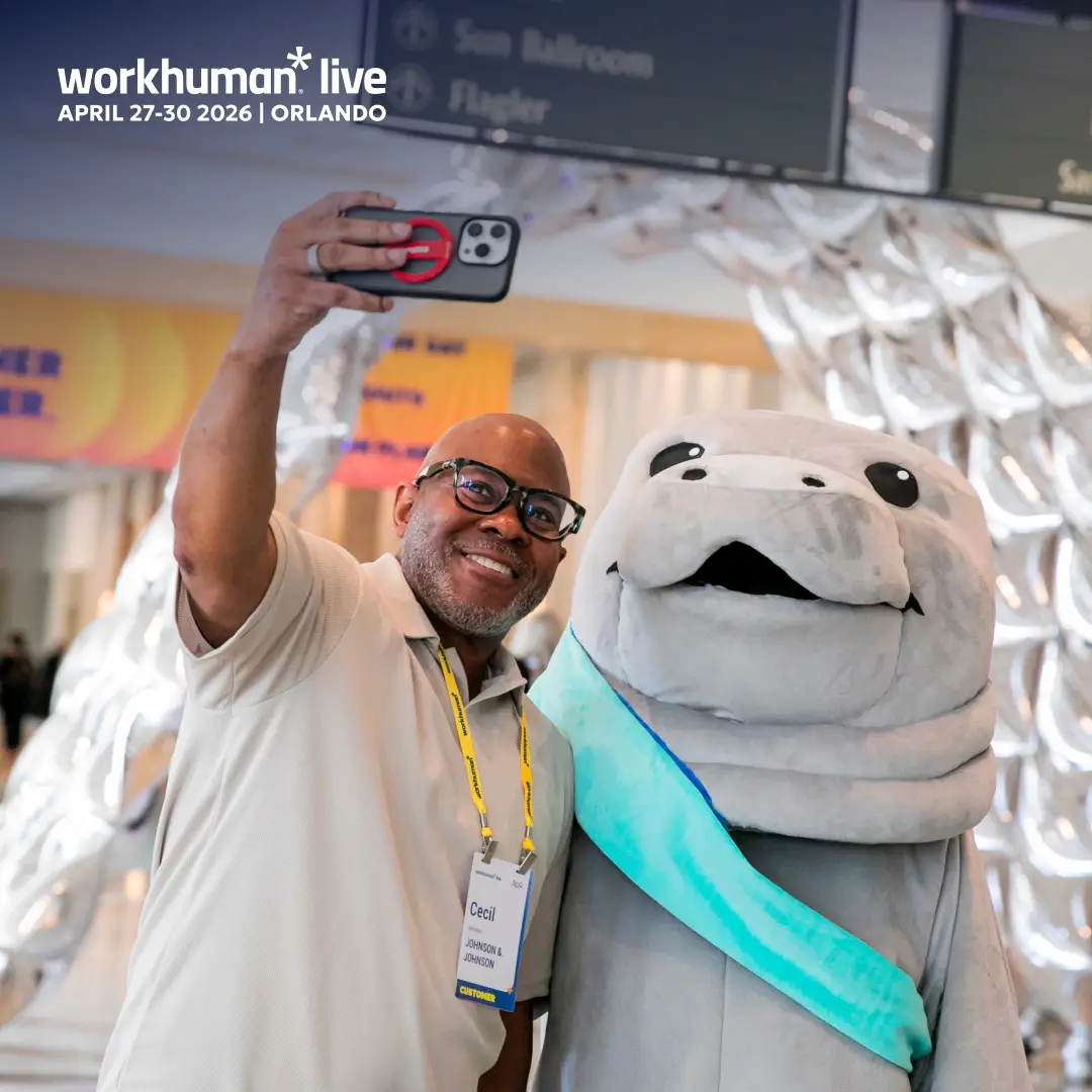 A man named Cecil is smiling while taking a selfie with a large, friendly mascot resembling a manatee. He is wearing glasses, a light grey shirt, and a name tag that reads "Cecil" with the company name Johnson & Johnson. In the background, there are colorful event banners and signs indicating directions, suggesting a lively atmosphere at the Workhuman Live conference taking place from April 27-30, 2026, in Orlando.