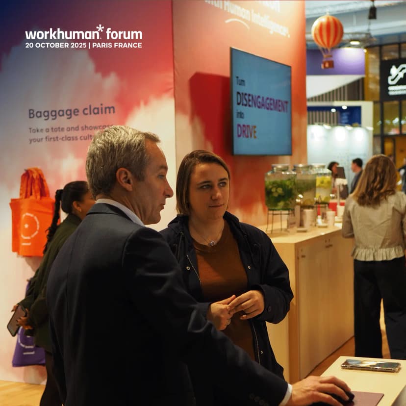 A professional networking moment at the Workhuman Forum in Paris on October 20, 2025, featuring two attendees engaged in conversation in the foreground. A man with short grey hair in a dark suit jacket and light blue shirt gestures while talking to a woman with dark brown hair wearing a dark blue jacket over a rust-colored top with a silver necklace. Behind them, event branding displays 'workhuman* forum' and '20 OCTOBER 2025 | PARIS FRANCE' with orange tote bags hanging nearby. A large screen shows 'Turn DISENGAGEMENT into DRIVE' in white and orange text. A beverage station features glass dispensers with infused water (cucumber and lemon), and other attendees are visible in the background. A decorative orange and white striped hot air balloon hangs from the ceiling, adding whimsical flair to the modern conference space.