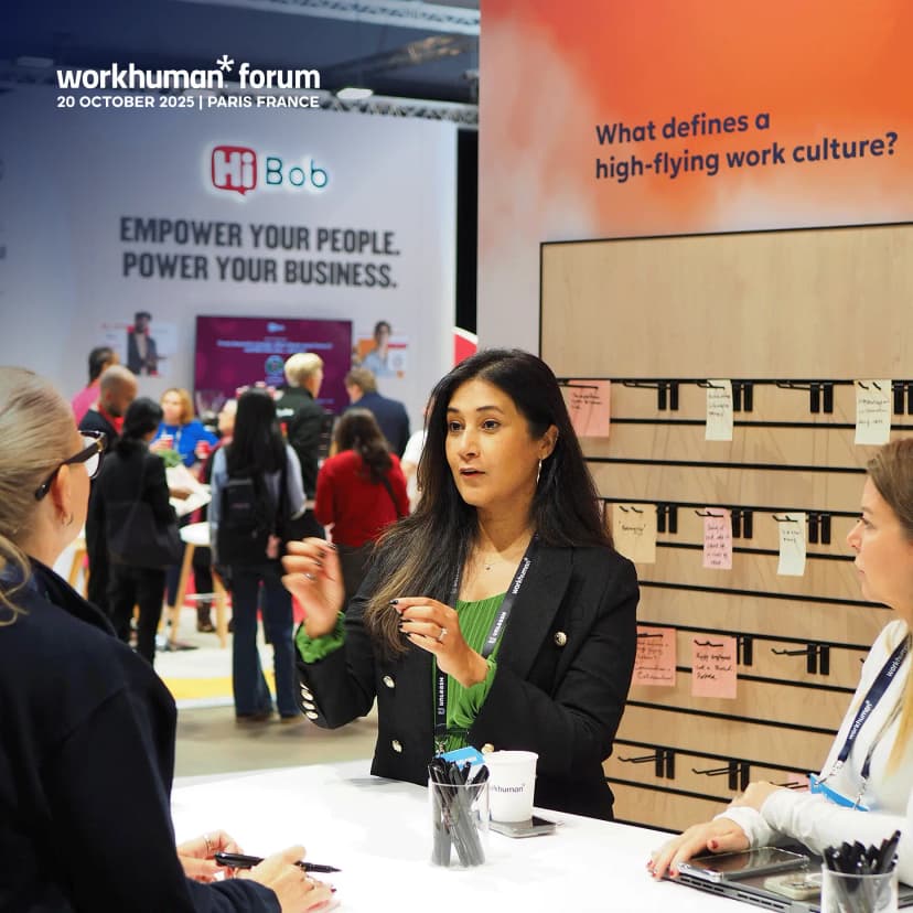 A woman with long dark hair is engaged in conversation at a Workhuman forum event, speaking to two attendees who are listening attentively. The backdrop features a banner with the text "What defines a high-flying work culture?" alongside another banner displaying "EMPOWER YOUR PEOPLE. POWER YOUR BUSINESS." There are various people mingling in the background, and the setting appears to be a bustling event with a modern, professional atmosphere.