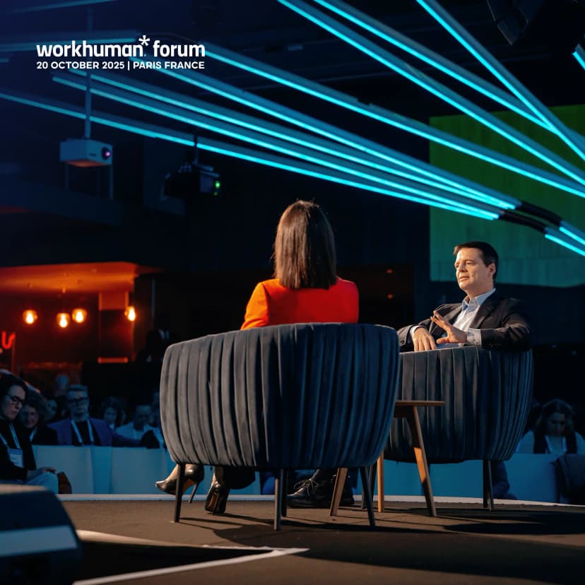A speaker at the Workhuman Forum sitting opposite another speaker, who is wearing a bright red outfit. The scene features modern-style chairs and vibrant blue lighting in the background, suggesting a professional event with an engaged audience in attendance. The top of the image displays the text "workhuman forum 20 OCTOBER 2025 | PARIS FRANCE."