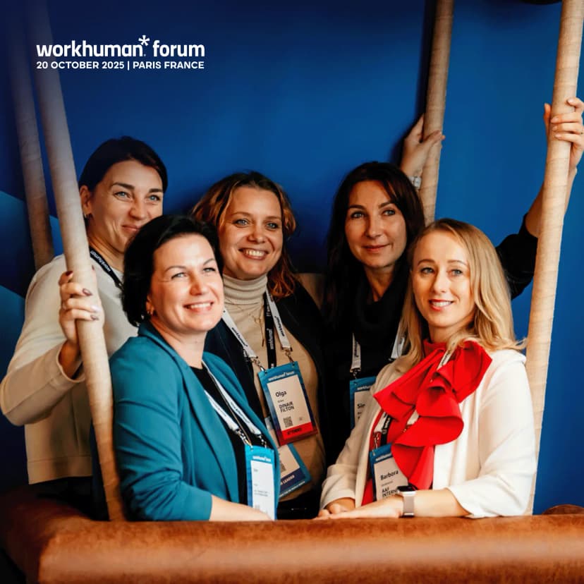 A group of five women smiling together in a booth setup at the Workhuman Forum in Paris, featuring a blue backdrop. Each woman is wearing a name tag, and they are positioned around a rustic-looking structure, engaging and enjoying the moment. The event details "20 OCTOBER 2025 | PARIS FRANCE" are visible at the top of the image.