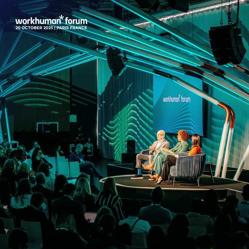 A panel discussion at the Workhuman Forum in Paris on October 20, 2025, featuring three panelists seated in modern dark grey armchairs on a circular stage. From left to right: an older man with glasses and grey hair in a light grey suit gesturing with his hands, a woman with curly reddish-brown hair wearing a green blazer and patterned dress, and a woman with straight dark hair in an orange top. Behind them, large screens display the 'workhuman* forum' logo against a teal background and abstract green wave patterns. The venue features striking teal light tubes crisscrossing the ceiling overhead, creating a futuristic ambiance, with black line array speakers suspended above. The audience sits in rows of white chairs in the foreground, dressed in dark clothing, some looking at the stage while others check phones or tablets.