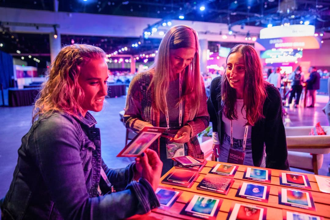 Three women are gathered around a table examining colorful cards at a lively conference setting. The atmosphere is bright and vibrant, with purple lighting, showcasing various interactive displays in the background. Each woman displays a sense of engagement and smiles as they interact with the cards.