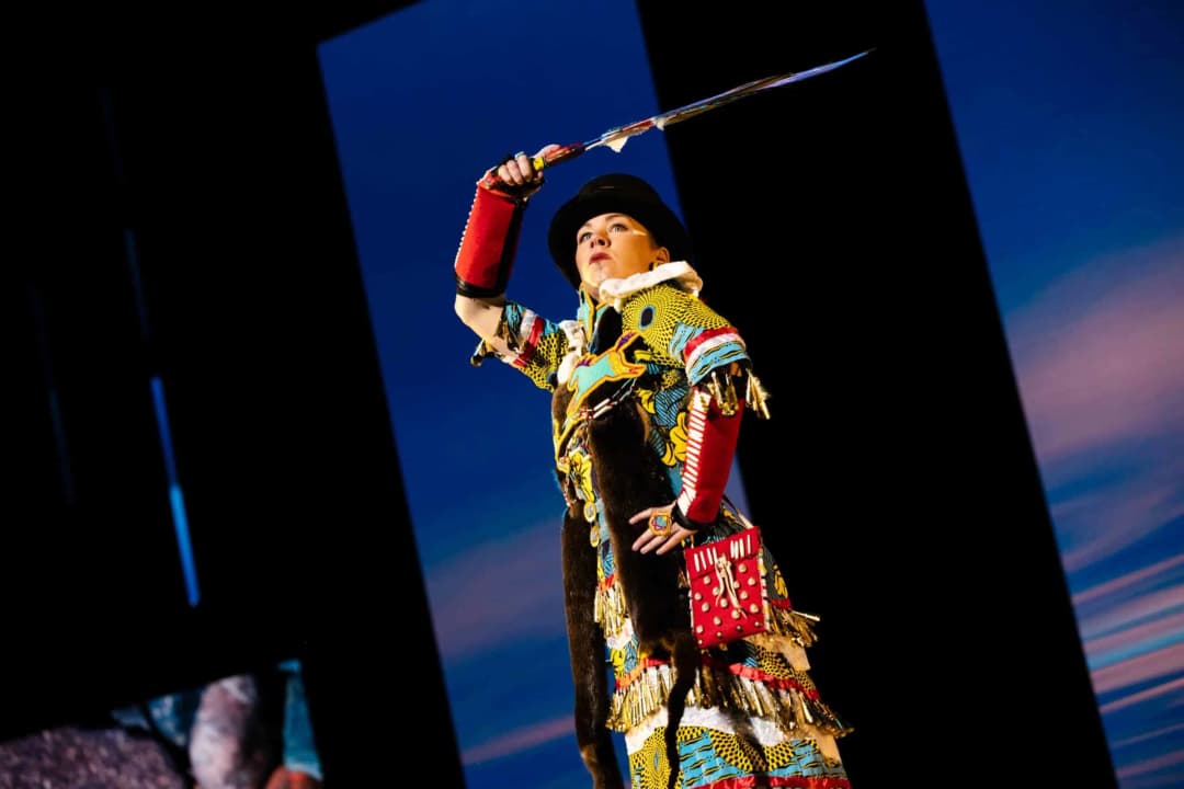 A performer stands confidently on stage, dressed in a vibrant, patterned outfit adorned with colorful fabrics, fringe, and accessories. She is holding a sword raised above her head, showcasing an expression of determination. The background features a scenic blue and purple gradient, enhancing the dramatic atmosphere of the performance.